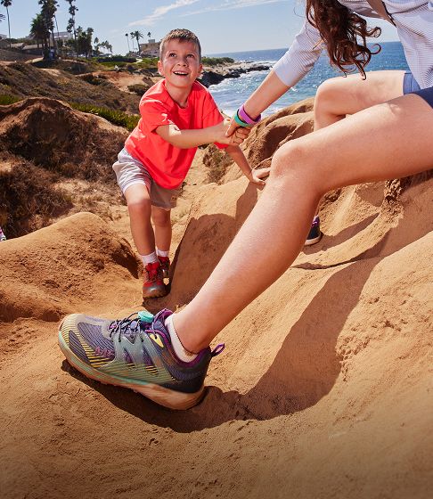 A mother and child playing together on a sand dune wearing Merrell shoes and apparel.